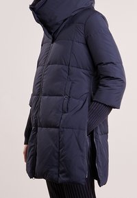 Person som bär en marinblå vadderad vinterjacka med hög krage, dragkedjestängning, sidofickor och ribbade stickade ärmar synliga under.