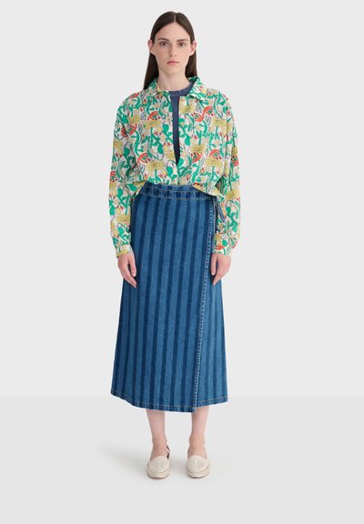 Femme portant une chemise à boutons à motifs floraux verts, jaunes et rouges, une jupe midi en denim bleu à rayures, et des chaussures blanches sans lacets.