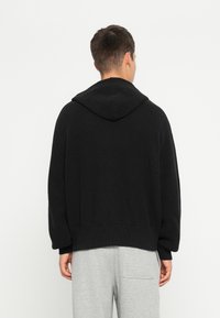 Svart strikket hettegenser med et teksturert mønster, løs passform, og en stor hette, kombinert med grå joggebukser med sidelommer.