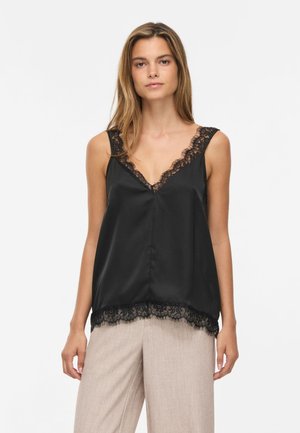 Femme portant un top sans manches en satin noir avec bordure en dentelle et un pantalon beige à jambes larges, debout devant un fond blanc uni.