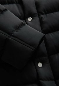 Detail černé péřové bundy s prošívaným designem, hladkým povrchem a kovovým drukem. Záběr na oblast rukávu.