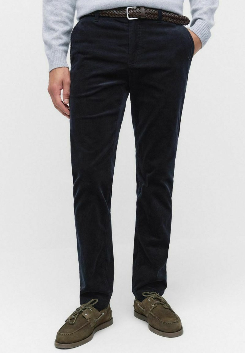 Marina blåa kordflossbyxor med rak passform och subtil textur, ihop med ett flätat brunt läderbälte och bruna mockaskor.