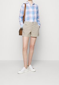 Osoba v modro-bílé kostkované košili, béžových šortkách, bílých teniskách a s hnědou kabelkou přes rameno stojící před jednobarevným pozadím.