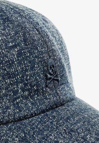 Gorra de denim de color azul oscuro con un acabado texturizado. Presenta un detalle de calavera y tibias negras bordadas en el lateral.