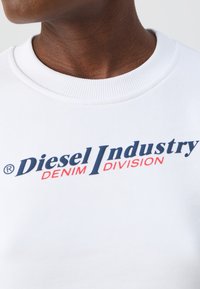 Biela mikina má rebrovaný okrúhly výstrih a výrazne zobrazuje "Diesel Industry" v námorníckej modrej farbe a "DENIM DIVISION" v červenom texte.
