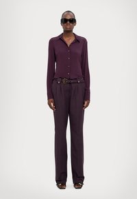 Camicia a maniche lunghe bordeaux con bottoni abbinata a pantaloni a gamba larga a vita alta. Gli occhiali da sole e una cintura decorativa completano l'outfit.