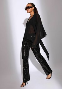 Fekete áttetsző felső fekete flitteres bővülő nadrággal, oversized napszemüveggel és pántos fekete magassarkúval kombinálva. Simaságot árasztó anyagok.