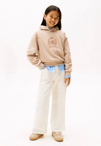 Jovem sorridente, usando capuz bege com logótipo de brasão, camisa azul às riscas, calças largas brancas e sapatos bege, com uma mão no bolso.