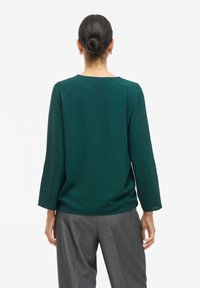 Grön texturerad långärmad topp med rund halsringning, ihopparad med grå skräddarsydda byxor. Tyget verkar mjukt och hänger löst.