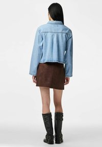 Moteris su ilgais tamsiais plaukais, vilkinti šviesiai mėlyną džinsinę striukę, rudą zomšinę mini sijoną ir juodus, iki kelių siekiančius batus su sagtimis, stovinti veidu nuošalyje.