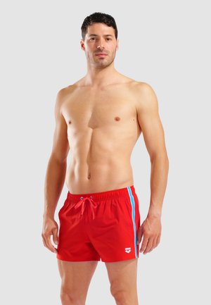 Uomo senza maglietta in piedi, che indossa pantaloncini da bagno rossi con strisce blu e bianche sui lati e una coulisse in vita.