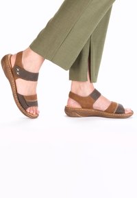 Brown and dark gray sandals with brown accents, featuring open toes, multiple straps, and a textured sole. Worn with green pants.
