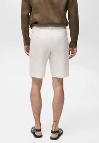 Pantaloni corti di un beige chiaro realizzati in lino, dotati di due tasche posteriori con chiusura a bottone. Indossati con una camicia a maniche lunghe marrone e sandali neri.