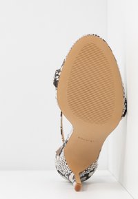 Semelle d'une chaussure à talons hauts avec semelle en caoutchouc beige et dessus à motif peau de serpent noir et blanc, inclinée contre un coin blanc.