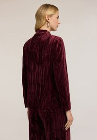 Burgundowa aksamitna marynarka z pionowymi fałdami, usztywnionym kołnierzem i długimi rękawami. Brak widocznych zapięć. Gładka tekstura podkreśla design.