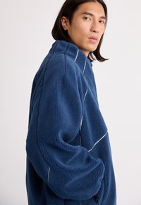 Navy fleece jacket with a high collar, featuring white stitching accents and oversized sleeves. Soft texture, casual design.