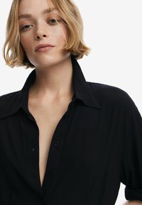 Chemise noire à manches courtes avec un col cranté, en tissu doux et coupe décontractée, dotée de boutons visibles sur le devant.
