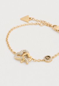 Χρυσό βραχιόλι με συνυφασμένες καρδιές, στολισμένο με διαυγή κρύσταλλα. Η αλυσίδα και το γούρι με λογότυπο συμπληρώνουν το σχέδιο.