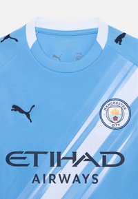 Šviesiai mėlynas futbolo marškinėliai iš tekstūruotos medžiagos, turintys baltas įstrižaines juostas, juodą Puma logotipą ir Manchester City ženklelį su detalėmis.