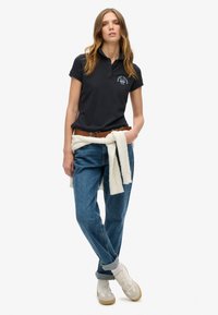 Polo de color marino con cuello, combinado con jeans de un azul claro y zapatillas blancas. Un suéter color crema atado a la cintura añade un detalle.
