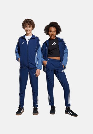Des survêtements de sport bleus avec des rayures blanches, fabriqués en tissu lisse. La veste présente une fermeture éclair et est portée par-dessus un crop top noir.