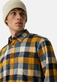 Geruite flanellen shirt in geel, marineblauw en wit met een button-down kraag en een borstzak. Heeft een gestructureerde stof en een crèmekleurige gebreide muts.