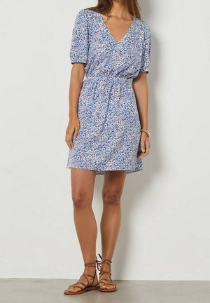 Femme portant une robe courte bleue à fleurs avec des manches bouffantes et des sandales plates beiges à lacets, debout contre un mur uni.