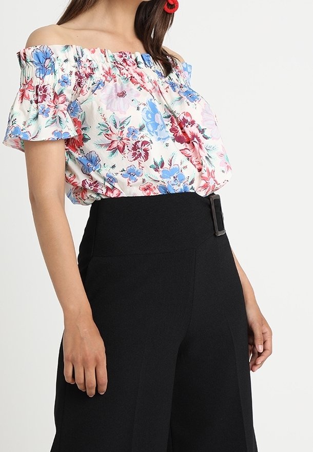 Donna che indossa una blusa off-shoulder con stampa floreale rossa, blu e rosa, abbinata a pantaloni neri a vita alta con una fibbia laterale.