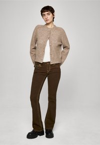 Hnědý pletený kardigan s kulatým výstřihem a předními knoflíky, kombinovaný s hnědými rozšířenými džínami s kontrastním prošitím a černými platformovými botami.