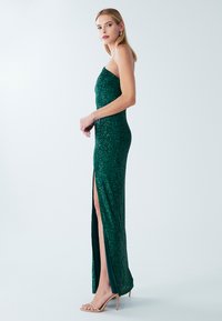 Žalia aplikacijomis puošta be petnešėlių suknelė su šoniniu skeltuku. Aptempta silueto, blizgaus tekstūros ir ilgo ilgio. Derinama su kūno spalvos aukštakulniais sandalais.