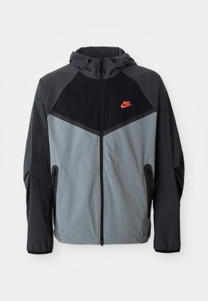Veste à capuche Nike présentant un schéma de couleurs noir et gris avec un design géométrique, des poches zippées et un logo rouge proéminent sur la poitrine.