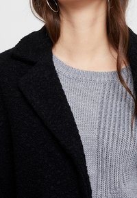 Czarny płaszcz o fakturze z klapami na jasnoszarym żeberkowanym swetrze. Widok z bliska na dekolt i szczegóły tkaniny.