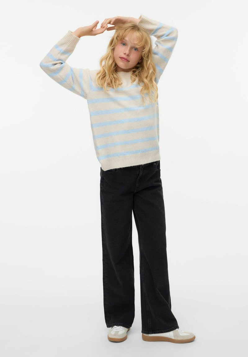 Pull en maille à rayures bleu clair et crème, pantalon large noir et baskets blanches avec semelles beiges. Mannequin pose avec les bras levés.