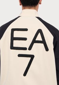 Pánská bunda s krémovým tělem a tmavě námořnickými rukávy, výrazné černě vyšívané "EA" a "7" na zadní straně, dekorativní ševová textura.