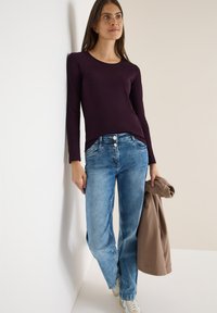 Langærmet, tætsiddende lilla top parret med lyseblå lige ben jeans. Modellen holder en beige sweater og står foran en neutral baggrund.
