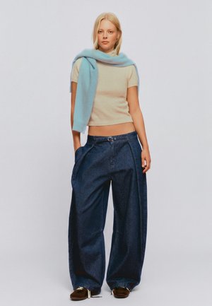 Jeune femme portant un top beige court, un pull bleu clair posé sur les épaules, un jean large bleu foncé et des chaussures marron.
