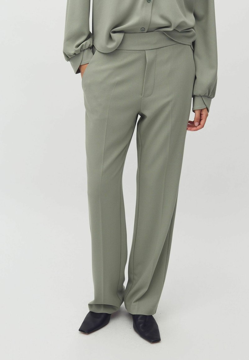 Pantalons ajustés vert clair avec une texture lisse, design à jambe droite et poches latérales, associés à des chaussures noires à bout pointu.
