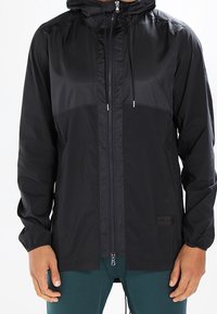 Black, lightweight jacket with a zip front, hood, and elastic cuffs. Features a smooth texture and contrasting upper section design.