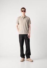 Beige polo-skjorte med krage, mørk denim-jeans og brune slip-on sko. Personen har mørke solbriller. Enkel bakgrunn.