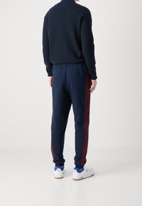 Marinblå joggingbyxor med vinröda sidoremsor, elastisk midjeresår och konisk benform. Kombinerade med vita sneakers med blå detaljer.