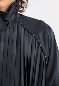 Black pleated jacket with a high collar, featuring a smooth texture and decorative silver hardware along the shoulder seam.