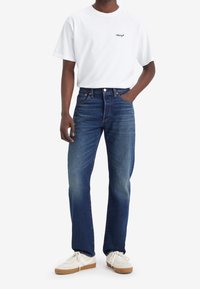 Vit kortärmad t-shirt med svart "Levi's"-logotyp, tillsammans med mörkblå raka jeans och vita sneakers med beige detaljer.