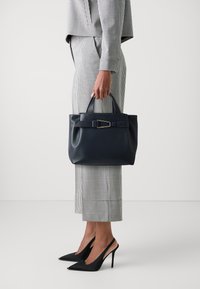 Námořnická kožená kabelka se strukturovaným designem, krátkými rukojetěmi, odnímatelným popruhem a zlatým detailem s přezkou, v kombinaci s kostkovaným oblekem.