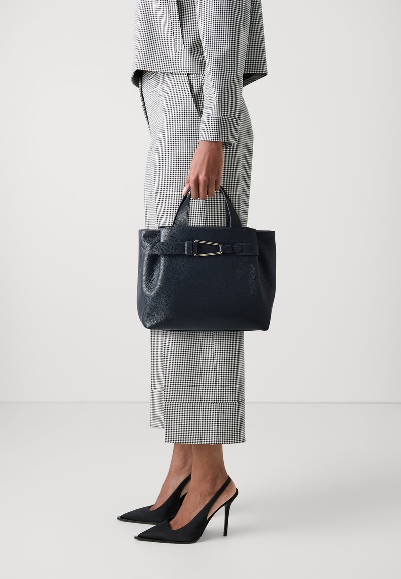 Námořnická kožená kabelka se strukturovaným designem, krátkými rukojetěmi, odnímatelným popruhem a zlatým detailem s přezkou, v kombinaci s kostkovaným oblekem.