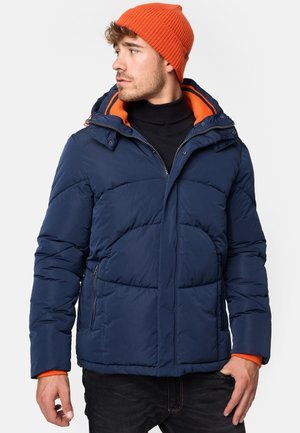 Navy puffer jacket with orange accents, featuring a hood, zippered pockets, and quilted texture. Worn over a black turtleneck. Orange beanie.