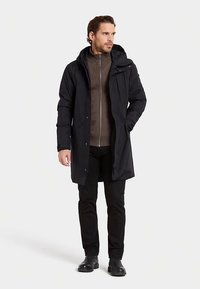 Svart vattentät parkas med luva, frontdragkedja och tryckknappsstängning, bärs över en grå zip-up-jacka. Kombineras med svarta byxor och stövlar.