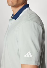 Light green polo shirt with a navy ribbed collar, white stripe detail, and white three-stripe logo on the left sleeve. Short sleeves.