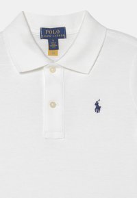 Vit pikétröja i bomull, med knappslå, platt krage och en marinblå broderad logotyp på vänster bröst.