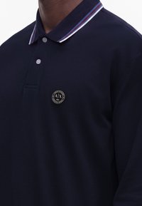 Homme portant un polo bleu marine avec deux boutons et un col rayé, affichant un logo rond Armani Exchange sur la poitrine.