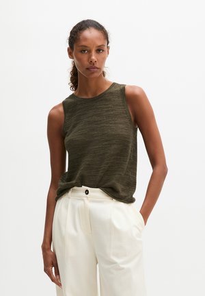 Femme portant un top en maille sans manches vert foncé et un pantalon blanc taille haute, une main dans la poche, sur un fond uni.